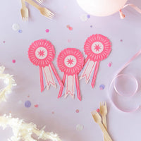 Three pink show ribbon napkins arranged on a lavender tabletop with pastel confetti and wooden forks—perfect for a pony-themed celebration.