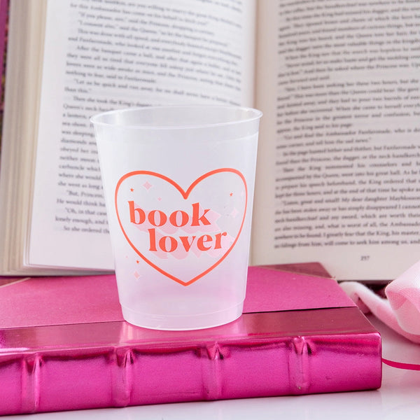 A clear flex cup printed with the phrase "book lover" in playful coral lettering surrounded by sparkly pink stars, placed on a shiny pink book with an open novel behind it.