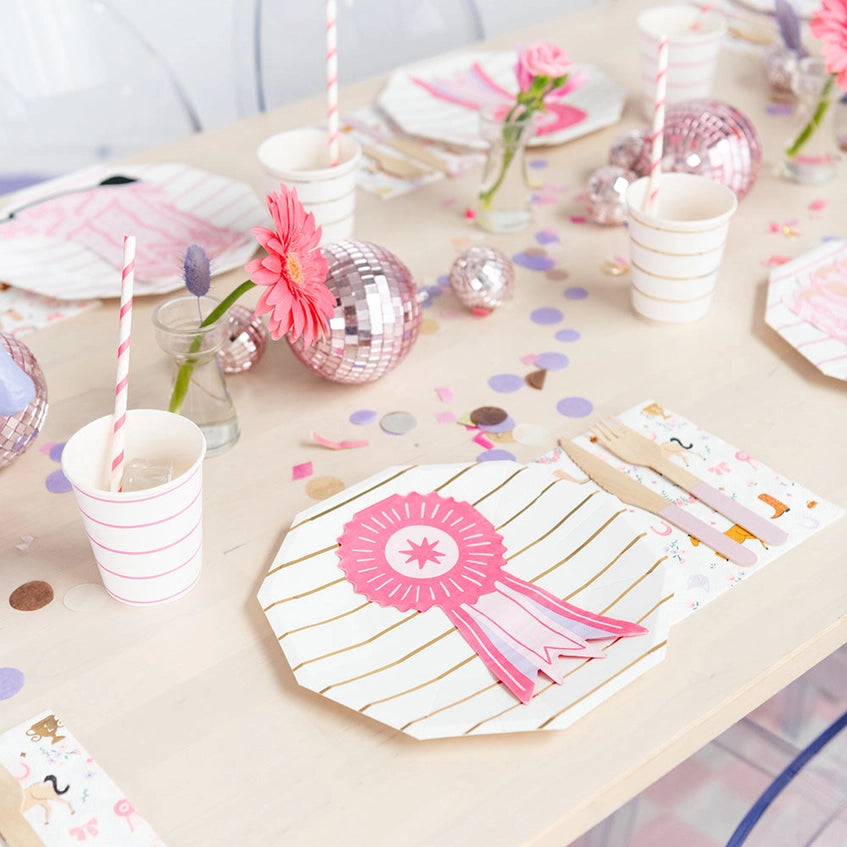 Angled shot of party table featuring pink striped cups, wooden forks, rhinestone straws, pink show ribbon napkins, and mini disco balls on a light wood surface.