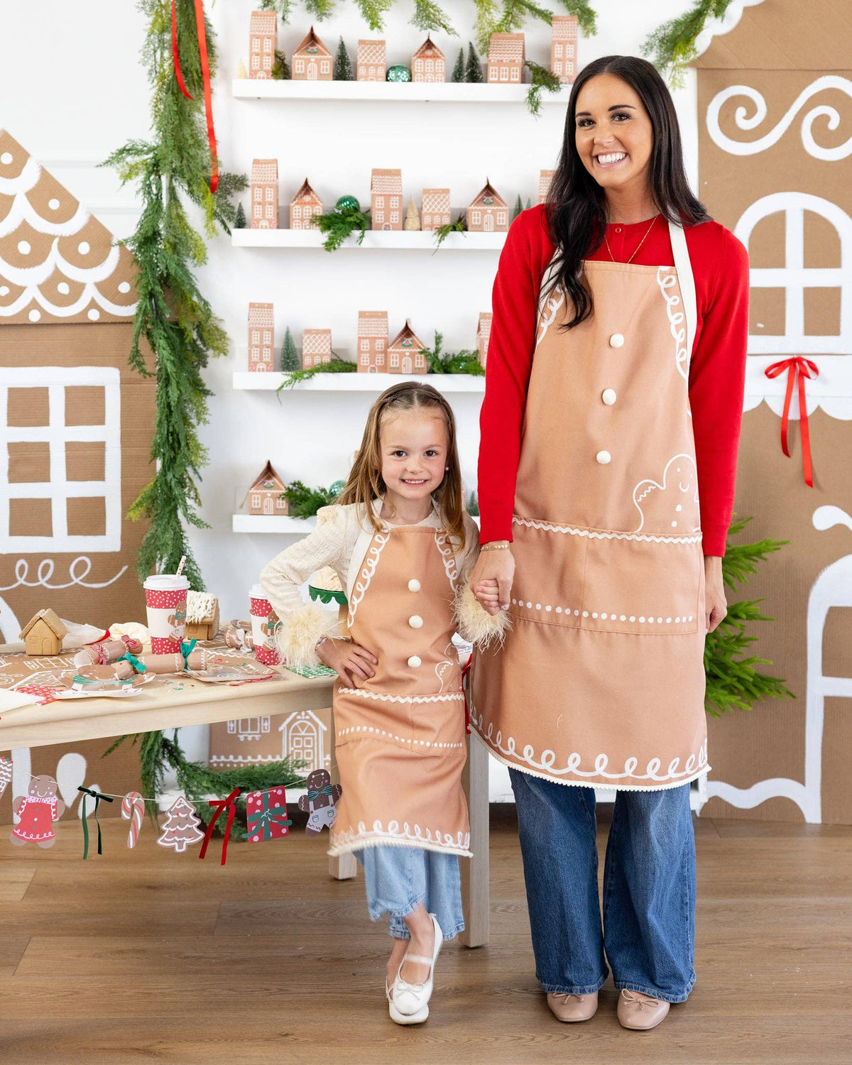 Mother and daughter smiling together wearing matching gingerbread aprons with white icing-style trim and pom-pom buttons.