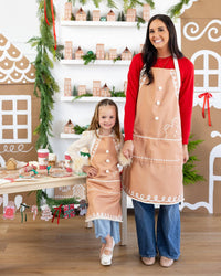 Mother and daughter smiling together wearing matching gingerbread aprons with white icing-style trim and pom-pom buttons.