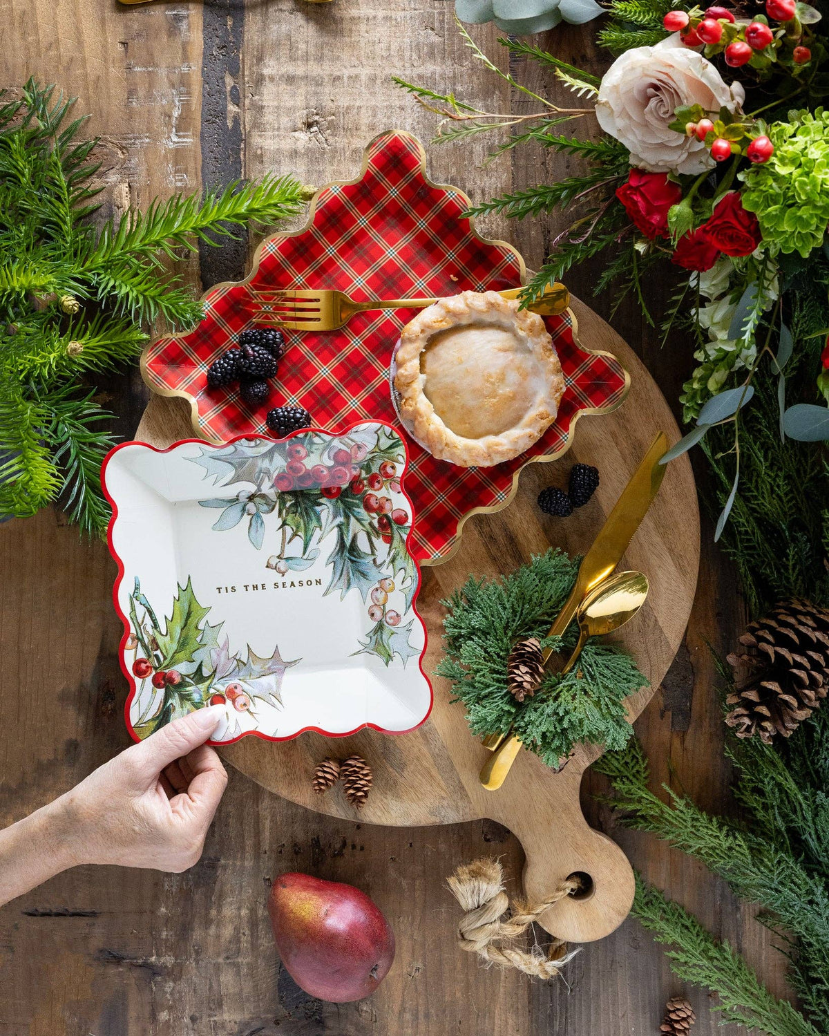 Hand holding holly plate beside mini pie and plaid plate on festive wooden setup