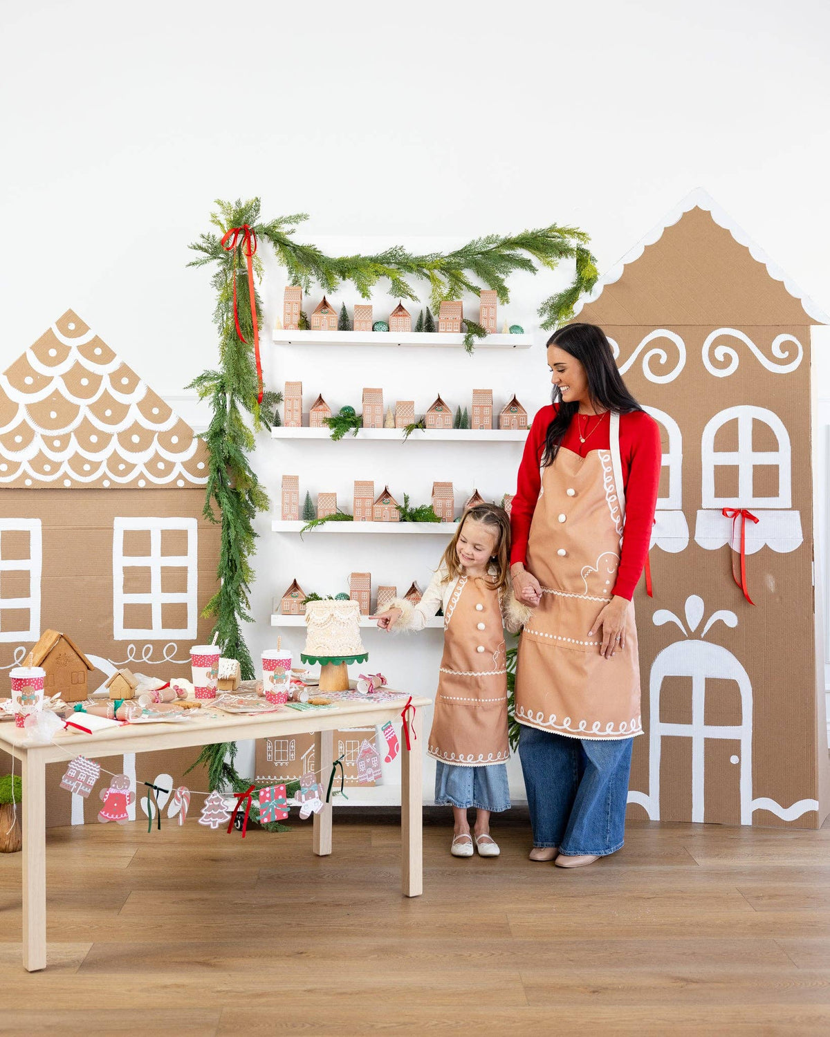 Mother and daughter smiling in matching gingerbread aprons surrounded by gingerbread house decorations and Christmas décor.