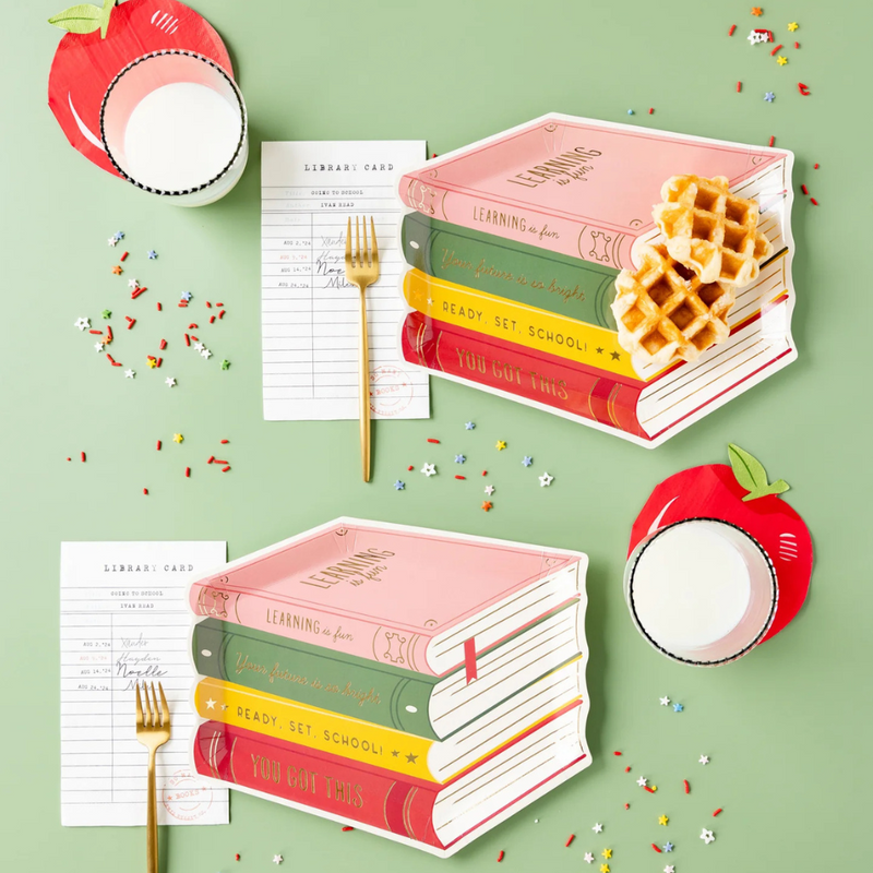 Styled table setting with book-shaped plates, apple cups, and library card napkins laid out on a mint green surface.