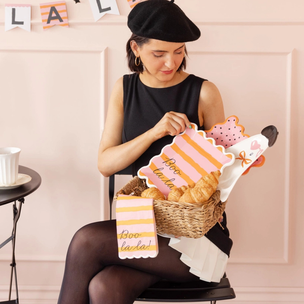 Woman holding a basket of Boo La La dinner napkins and matching plates while seated at a pink-walled table.