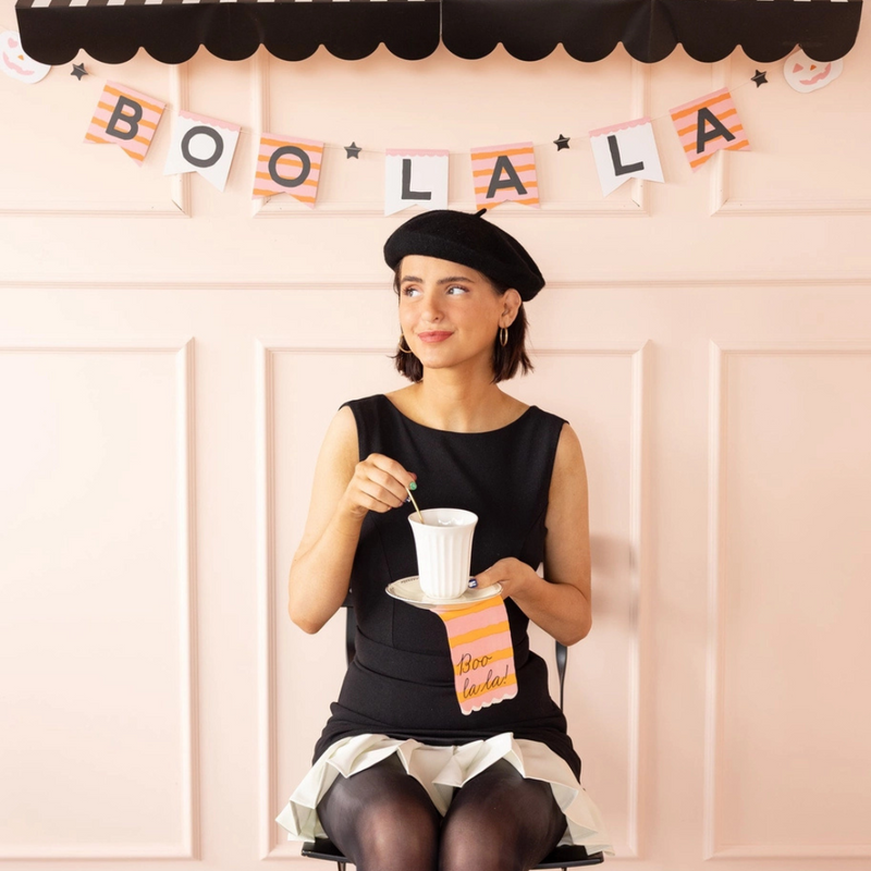 Woman stirring tea with a Boo La La napkin on her lap, seated in front of the coordinating Halloween banner.