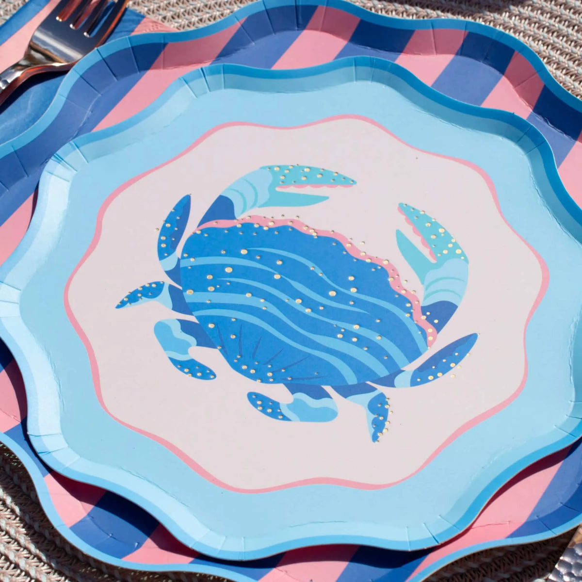 Close-up of the blue crab scalloped salad plate layered over a pink-and-blue diagonal-stripe plate on a woven mat, gold foil crab detail and scalloped edges in focus, flatware at the upper left. Angled detail shot.