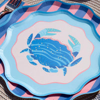 Close-up of the blue crab scalloped salad plate layered over a pink-and-blue diagonal-stripe plate on a woven mat, gold foil crab detail and scalloped edges in focus, flatware at the upper left. Angled detail shot.
