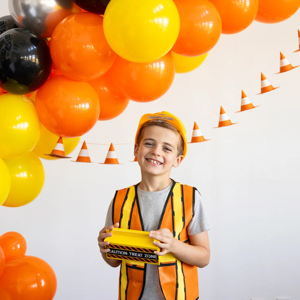 Boy in construction outfit holding treat box in front of cone banner and balloons