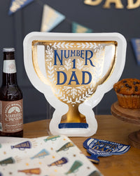 A close-up of the Number 1 Dad Trophy Plate next to a bottle of craft soda and a muffin, with Father's Day decor in the background.
