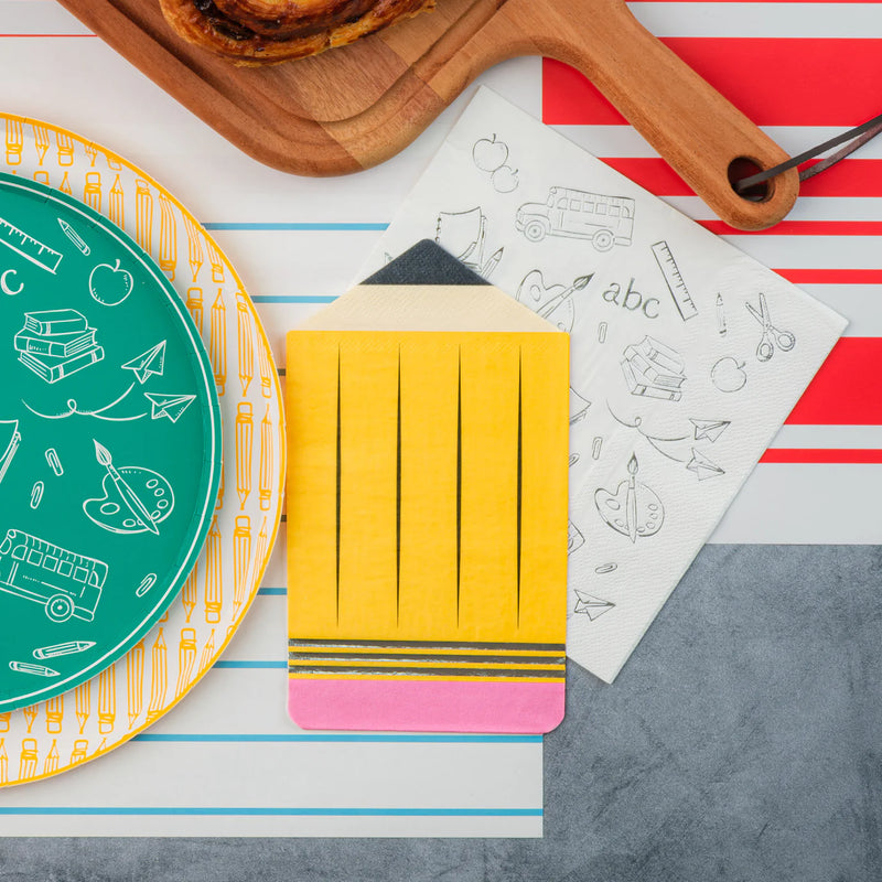 Close‑up of teal icon plate on notebook placemat next to yellow pencil napkin and wood board with pastries