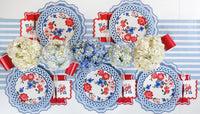 Wide overhead shot of a full table set for six with blue striped runner and white surface. Every place setting uses a dusty blue scalloped placemat under a white lattice-cut round charger and a red, pink, and blue floral scalloped paper plate. Red paper cup pouches roll-wrapped at each setting. White, blue, and cream hydrangea centerpieces.