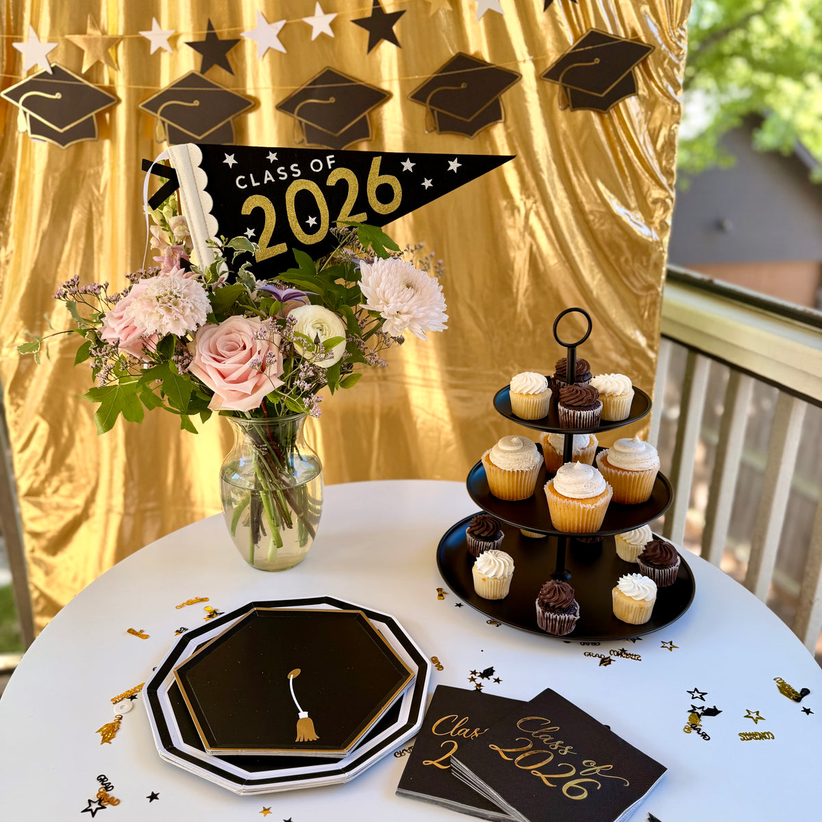 Class of 2026 graduation party tablescape featuring a black and gold felt pennant flag centerpiece, grad cap paper plates, foiled cocktail napkins, a three-tier cupcake stand, and a grad cap and star banner backdrop