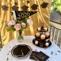 Class of 2026 graduation party tablescape featuring a black and gold felt pennant flag centerpiece, grad cap paper plates, foiled cocktail napkins, a three-tier cupcake stand, and a grad cap and star banner backdrop