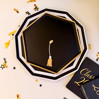Black and gold graduation cap shaped paper plates stacked in two sizes, styled with gold and black grad confetti on a white surface