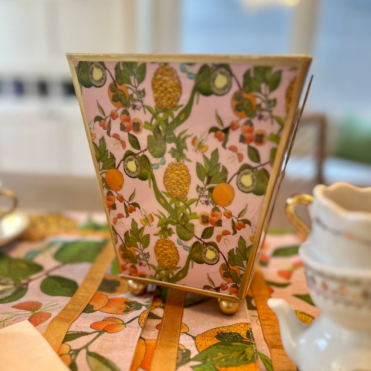 Make a statement with our Enameled Square Cachepot Planter, a playful yet practical table centerpiece. Decked out in a vibrant palette and a fruity motif of pineapples, limes, and oranges, this cachepot is trimmed in gold for that extra sparkle. Whether you're looking to brighten up your office desk, kitchen counter, or dining table, it offers a splash of spring and summer all year round. Versatile and visually stunning, it’s the centerpiece that brings your tablescape ideas to life.