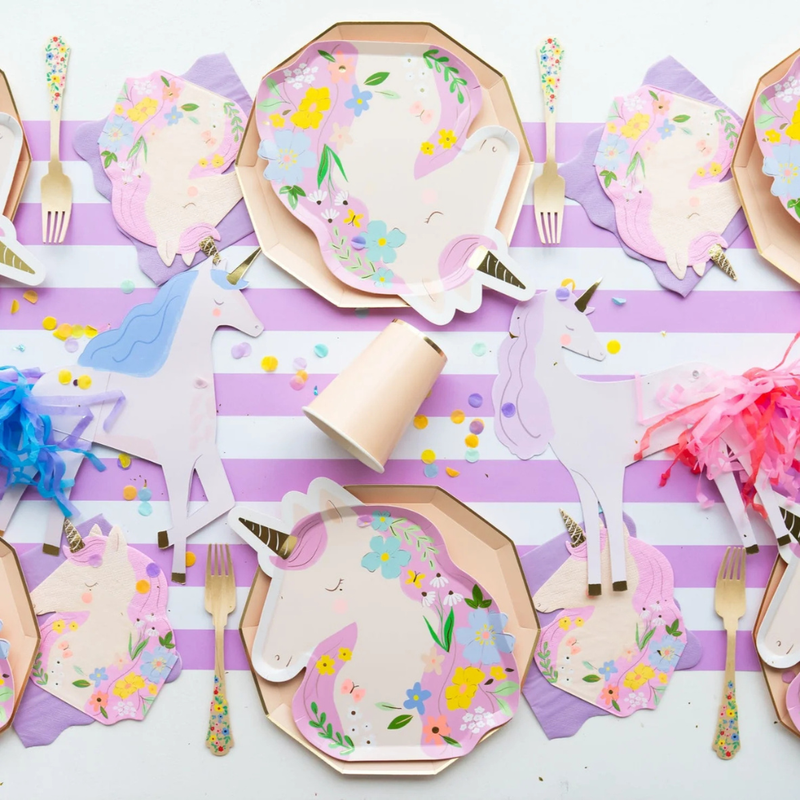 A festive unicorn-themed table setting featuring pastel-colored unicorn plates, cups, napkins, and confetti arranged on a lilac and white striped table runner.