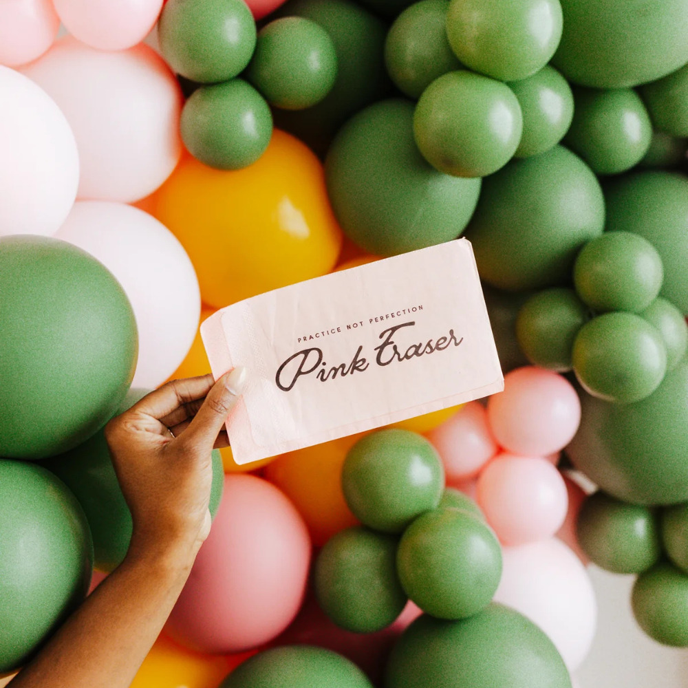 Hand holding a pink “Practice Not Perfection” eraser napkin in front of a colorful balloon backdrop.