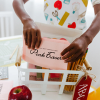 Close-up of a child holding a pink eraser napkin from a white basket filled with pencils and other school-themed supplies.