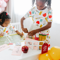 Two young girls in school supply print outfits engaging with a back to school party setup, including pink eraser napkins and activity placemats.
