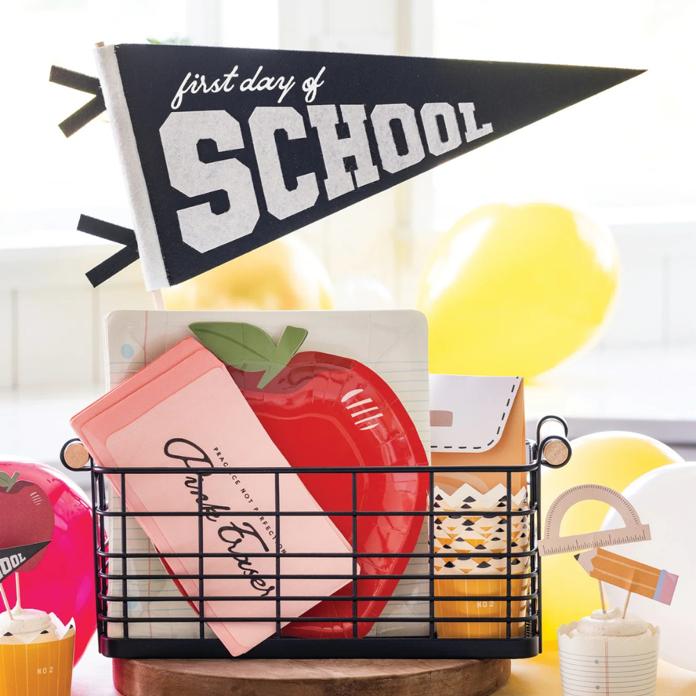 A black basket filled with school-themed party supplies, including pink eraser napkins and an apple-shaped plate, with a "First Day of School" pennant in the background.