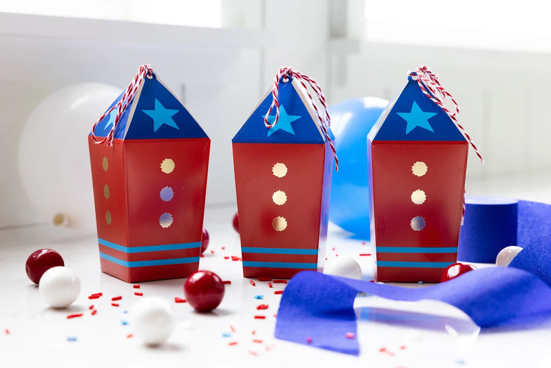 Set of three red, white, and blue rocket-themed treat boxes arranged with party decorations and sweets.