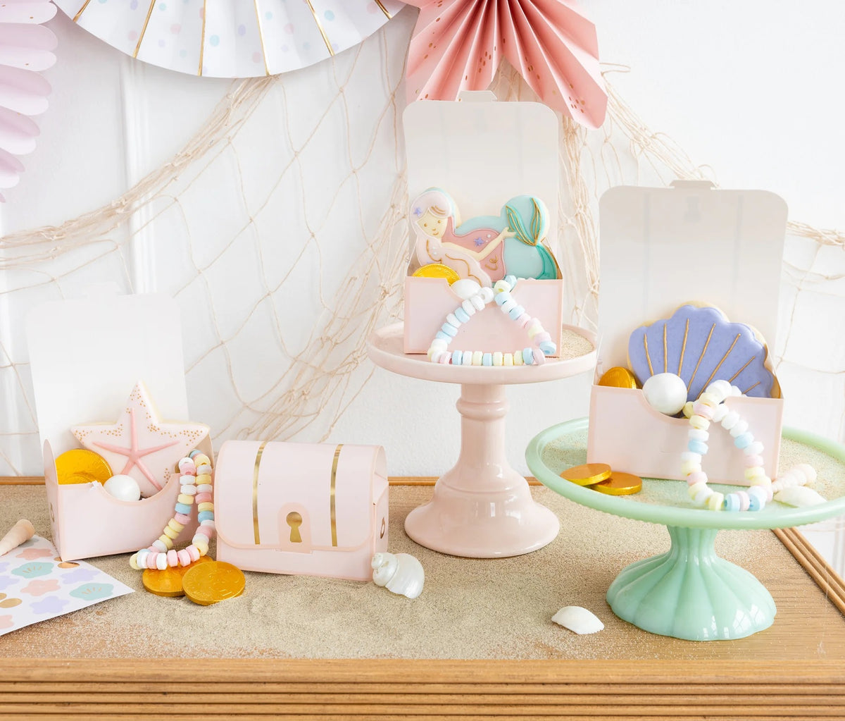 Adorable treasure boxes displayed on a beach-inspired table setting, complete with pastel-colored paper fan decorations and a charming fishing net backdrop.