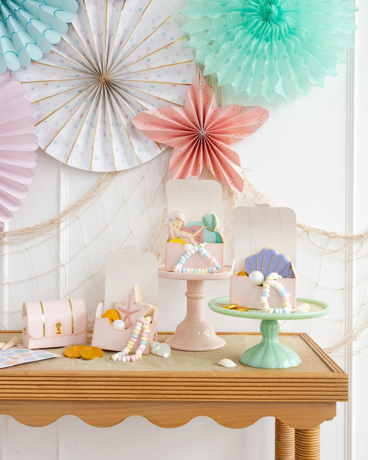 Beautifully styled mermaid treasure boxes on a sandy surface, brimming with candy jewelry, gold chocolate coins, and a sugar cookie shaped like a starfish.
