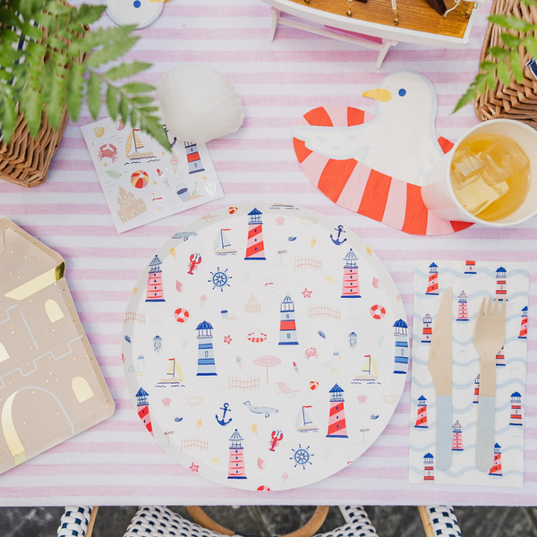 Overhead view of a seaside-themed table setting featuring lighthouse plates, wave-patterned napkins with cutlery, a seagull-shaped napkin, a cup of iced tea, seashell decor, and a sticker sheet with coastal illustrations.