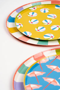 A set of dessert plates with colorful borders, stacked on charger plates, featuring summer-inspired designs of umbrellas and beach balls, perfect for a tropical or poolside party.