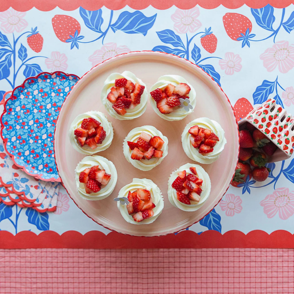 A top-down view of a dessert display with several cupcakes topped with whipped cream and chopped strawberries, arranged on a pink cake stand over a strawberry-patterned table runner with blue leaves and red scalloped edges.