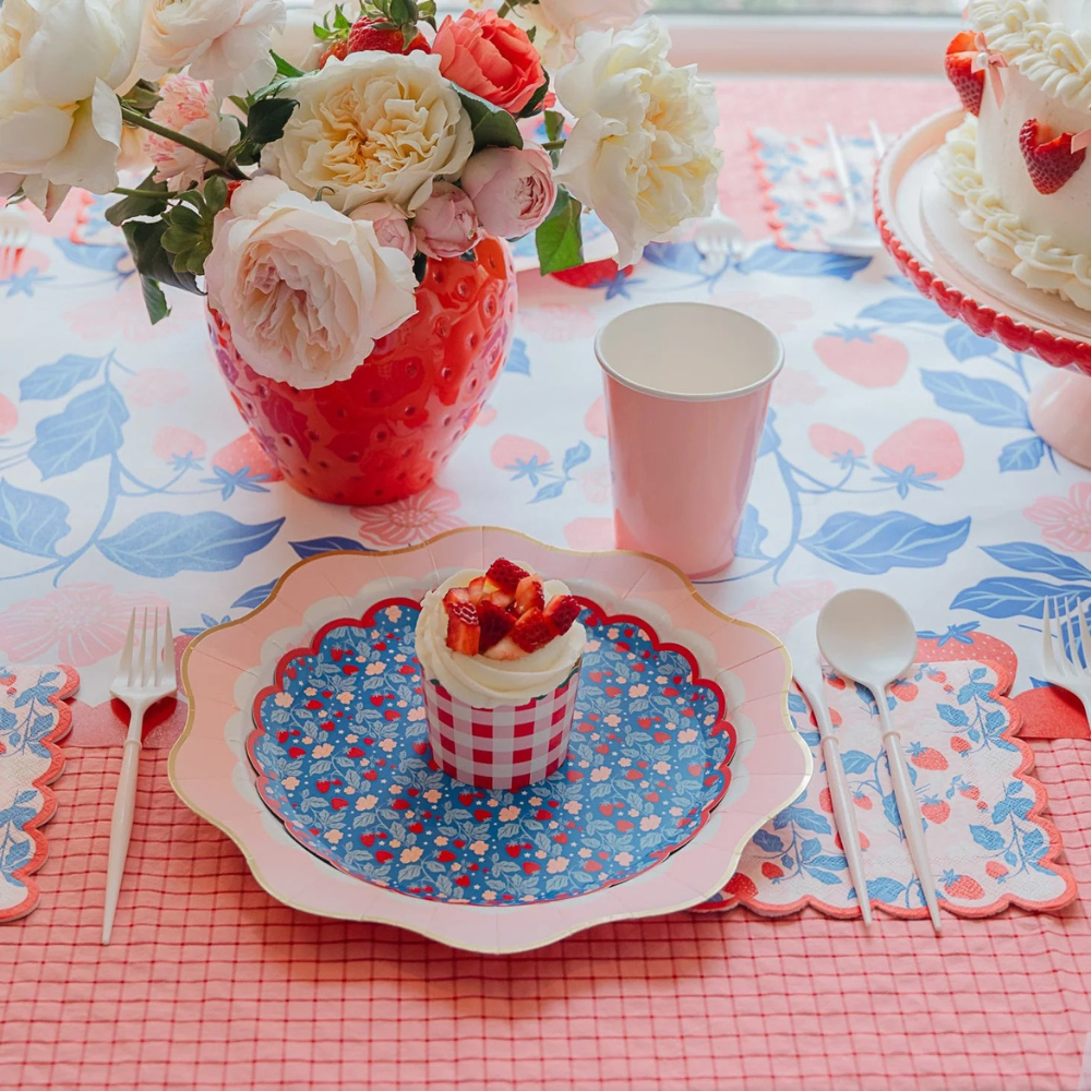 A colorful table setting featuring a floral centerpiece in a red strawberry-shaped vase, a pink and blue strawberry-patterned table runner, and a cupcake topped with strawberries on a decorative blue and pink scalloped plate.