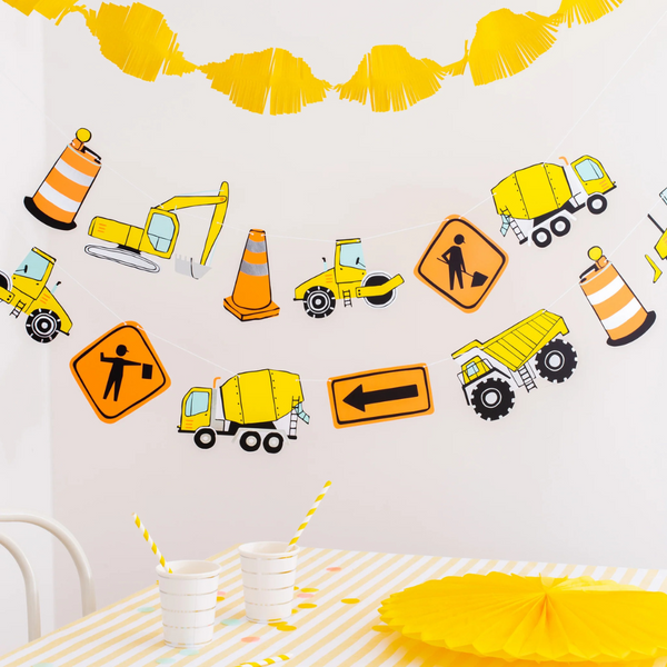A festive party setup with yellow garlands and a hanging construction-themed garland featuring signs, trucks, cones, and construction equipment.