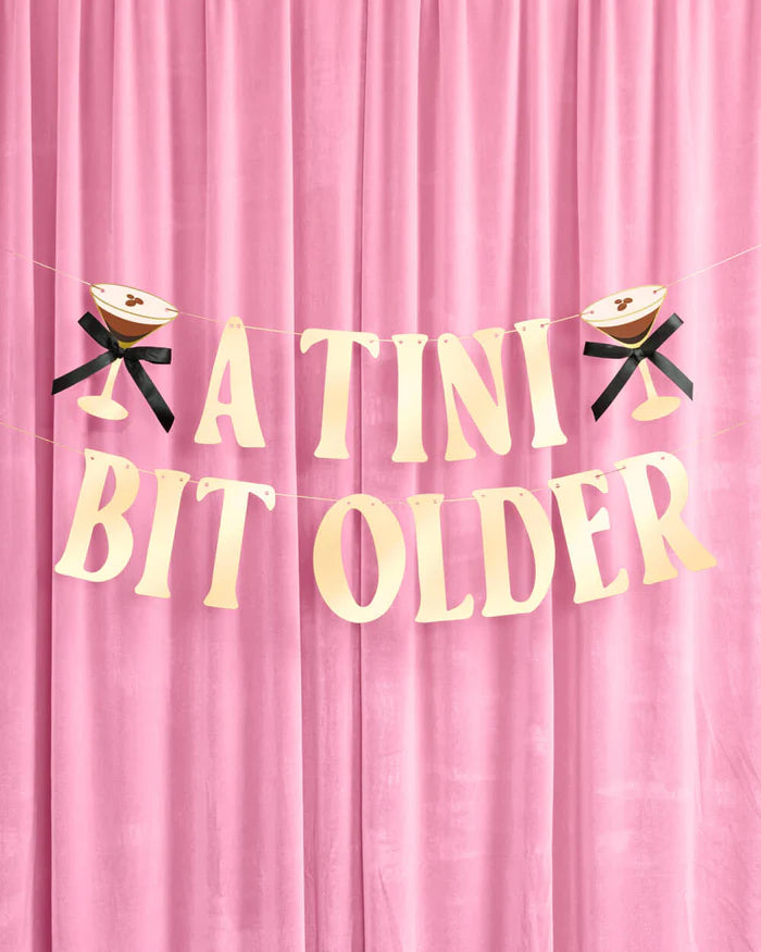 Full "A Tini Bit Older" gold foil banner pre-strung on two rows with espresso martini icons on either end against pink velvet curtain