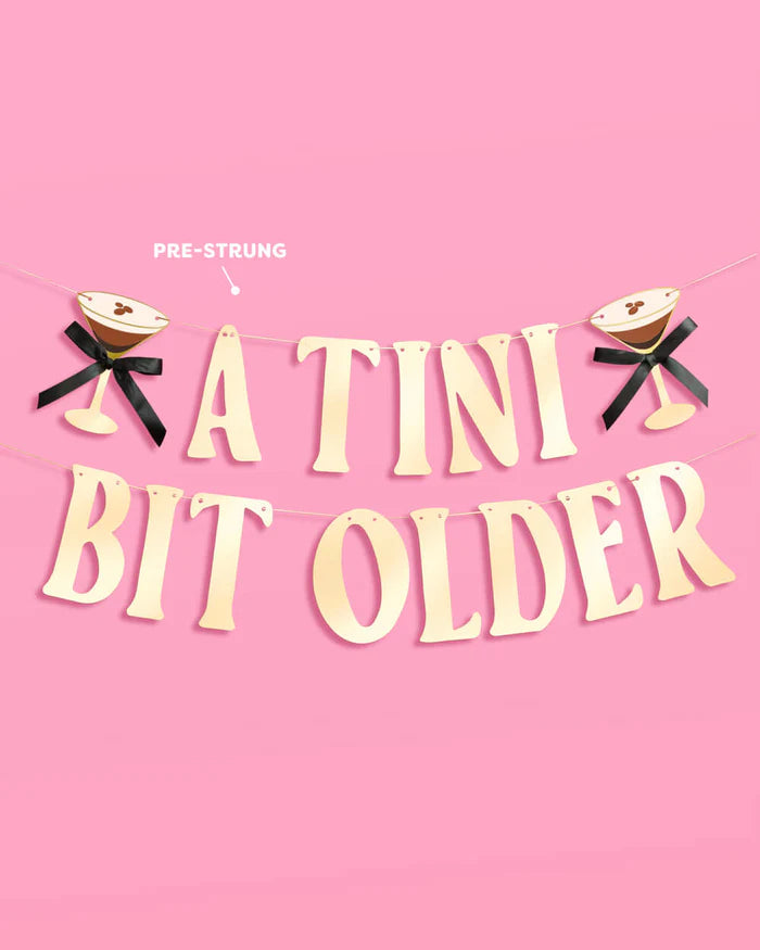 Pre-strung "A Tini Bit Older" two-tier gold foil birthday banner with martini glass icons on pink background, labeled "pre-strung"