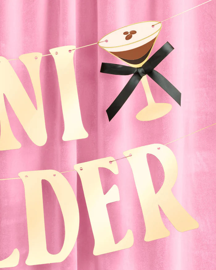 Close-up of gold foil "A Tini Bit Older" birthday banner letters with espresso martini glass icon and black bow hanging against pink velvet backdrop