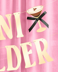 Close-up of gold foil "A Tini Bit Older" birthday banner letters with espresso martini glass icon and black bow hanging against pink velvet backdrop