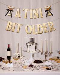 Styled espresso martini birthday table with "A Tini Bit Older" banner hanging above cake, candles, wine, martini glasses, cherries, and black bow place cards