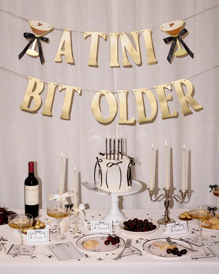 Styled espresso martini birthday table with "A Tini Bit Older" banner hanging above cake, candles, wine, martini glasses, cherries, and black bow place cards