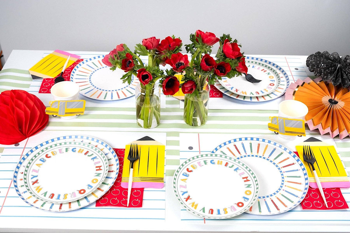 Back-to-school tablescape with alphabet paper plates layered under pencil-rim plates, yellow school bus napkins, pencil-shaped yellow napkins, red apple-print runner, and red anemone arrangements in glass vases