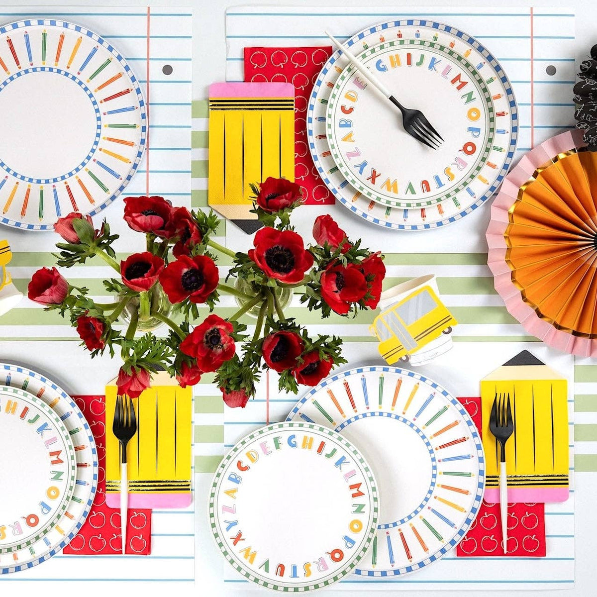 Angled tablescape view with alphabet plates, pencil-rim plates, yellow pencil-shaped napkins, school bus napkins, and red anemones on a green and white striped runner