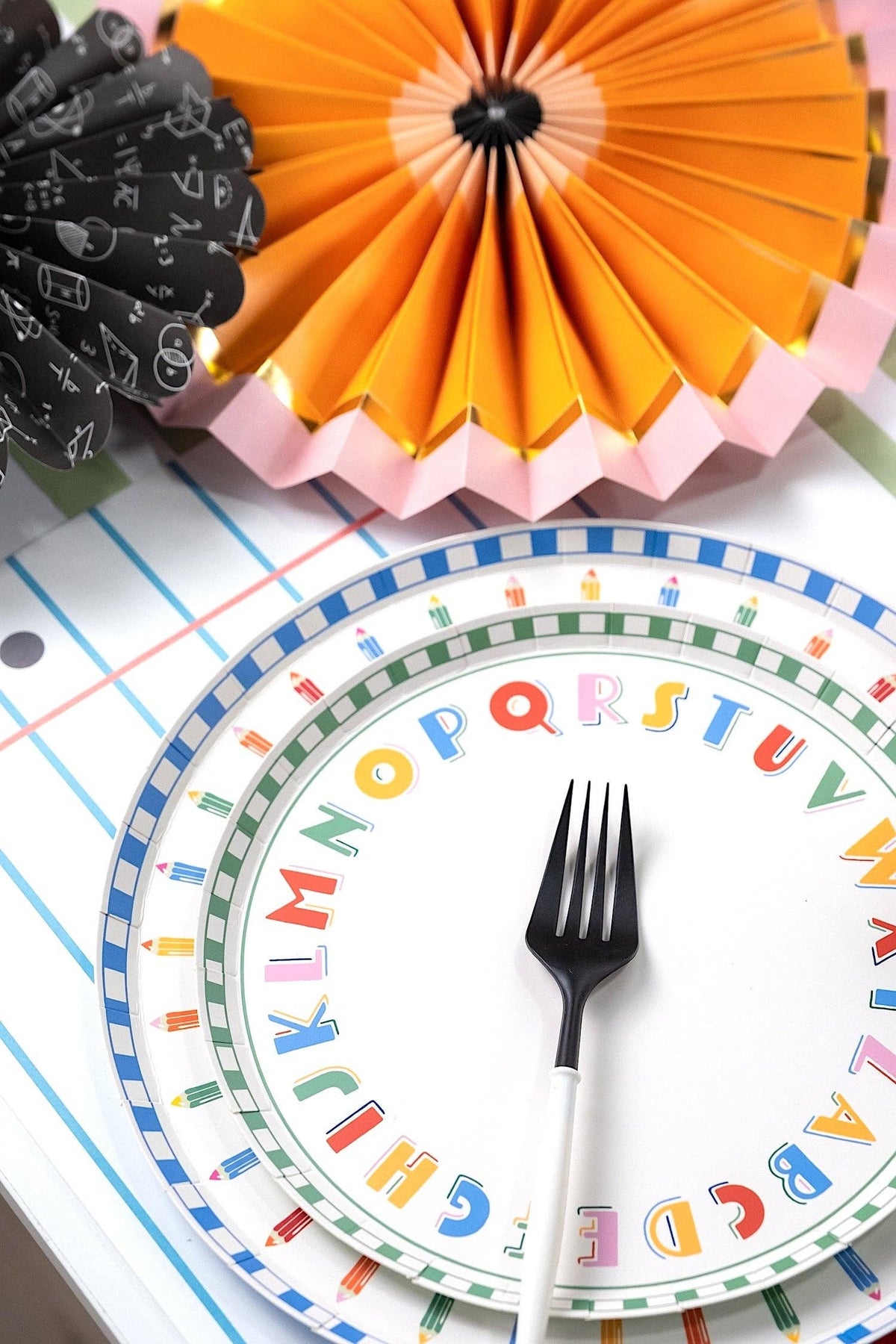 Close-up of an ABC alphabet paper plate layered under a pencil-rim paper plate on a notebook-paper-print table runner with an orange and pink paper fan and a black-and-white chalkboard-print fan in the background
