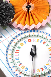 Close-up of an ABC alphabet paper plate layered under a pencil-rim paper plate on a notebook-paper-print table runner with an orange and pink paper fan and a black-and-white chalkboard-print fan in the background
