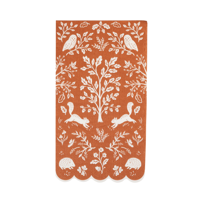 Overhead view of a folded rectangular terracotta paper dinner napkin featuring a white folk art woodland pattern and a scalloped bottom edge.