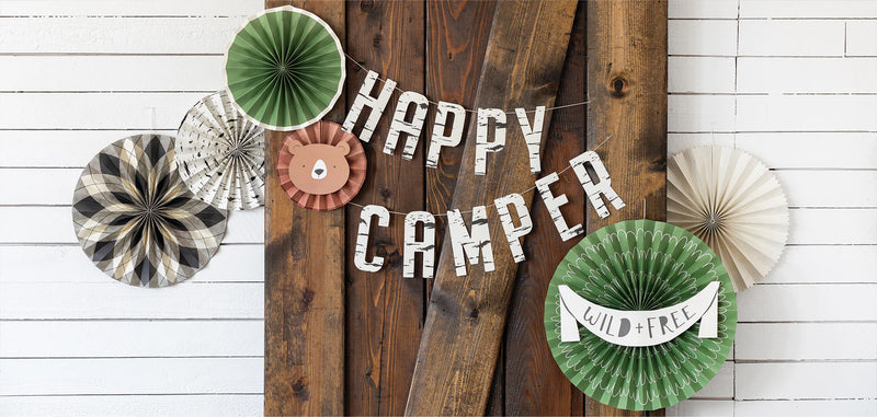 Close-up of hanging paper fans with “Happy Camper” banner and cute bear faceplate on wooden barn doors, styled for a camping-themed party.