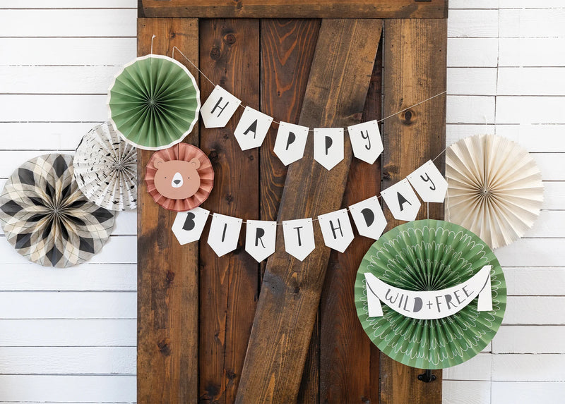 “Happy Birthday” banner hanging on wooden barn doors surrounded by green, plaid, and kraft paper fans for a cozy camping birthday party vibe.