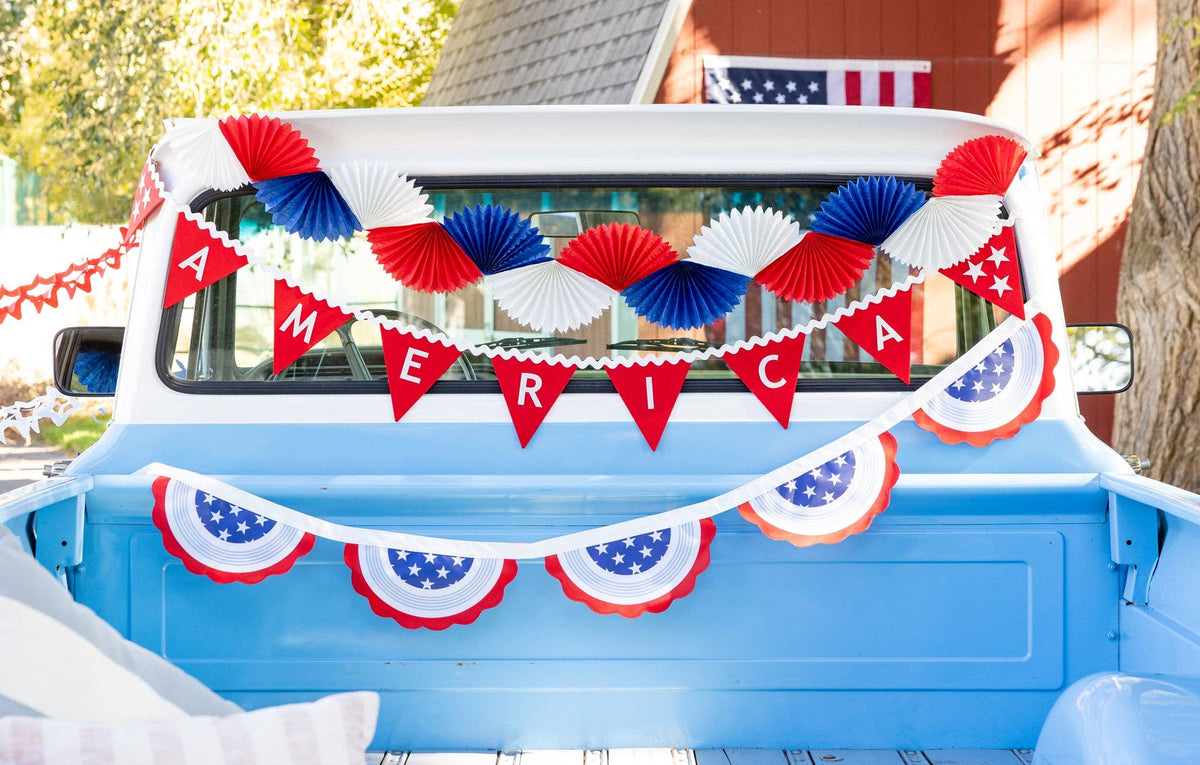Detail shot of America felt pennant banner with red white and blue paper fan garland hung across back window of blue vintage truck