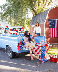 America felt pennant banner hung on vintage blue truck with two women and a dog sitting in the truck bed surrounded by patriotic party decor