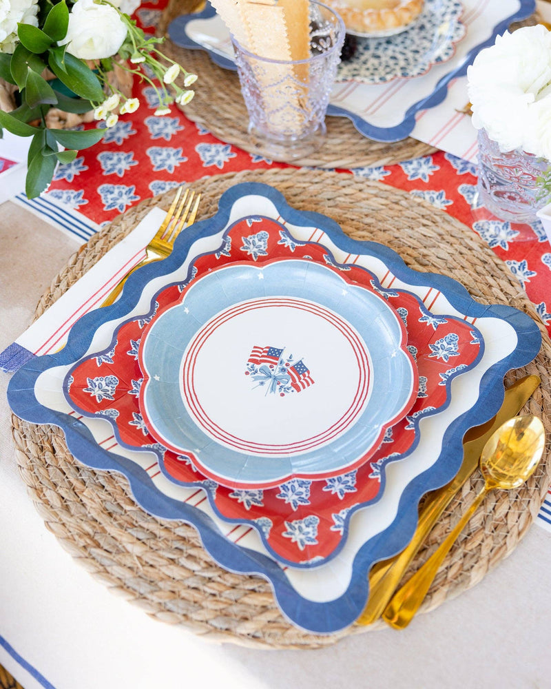 American flag scalloped paper plate layered on red block-print plate and blue scalloped striped charger on wicker placemat with gold cutlery — close-up of full Hamptons plate stack