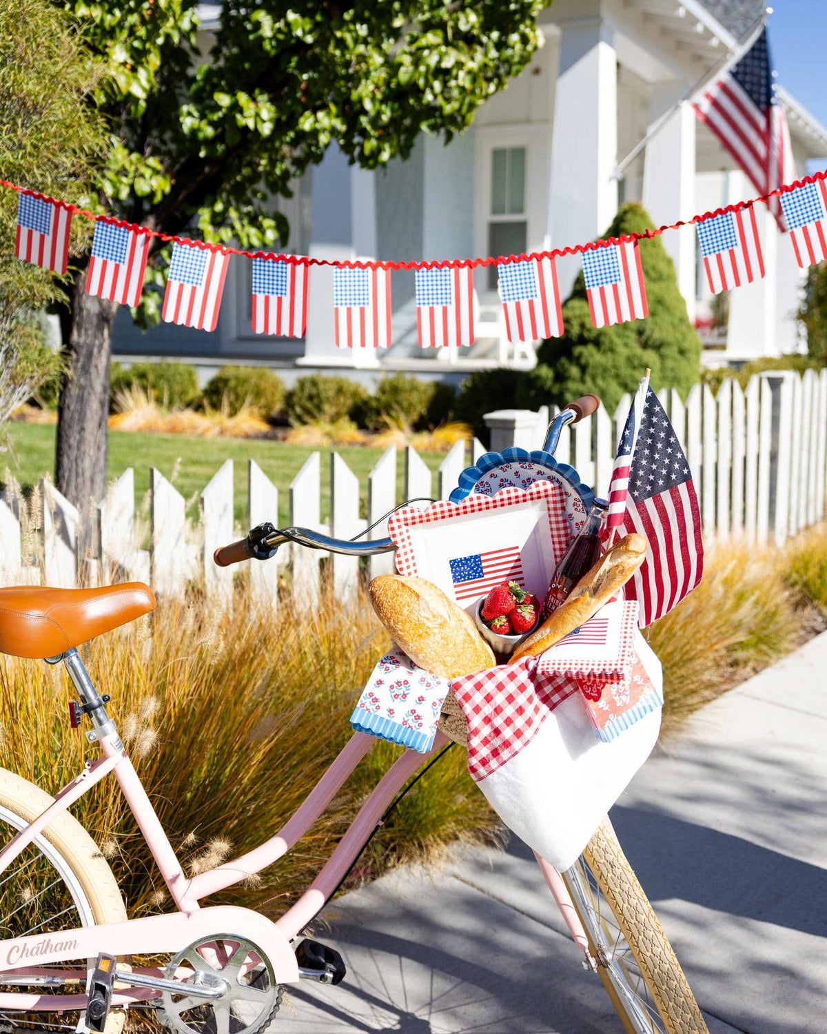 Pink bicycle with basket filled with gingham flag plates, floral scalloped plates, floral napkins, and American flags in front of white picket fence